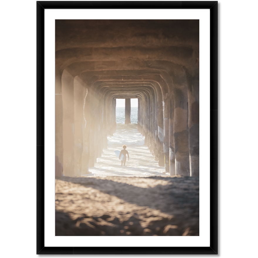 image of a surfer under a pier walking out into the waves carrying a surfboard
