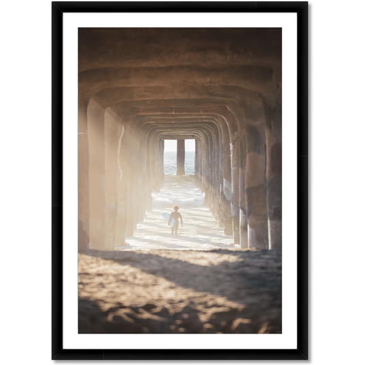 image of a surfer under a pier walking out into the waves carrying a surfboard