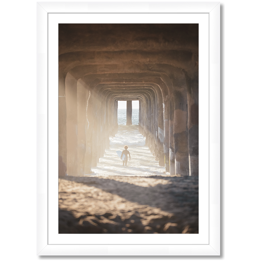 image of a surfer under a pier walking out into the waves carrying a surfboard