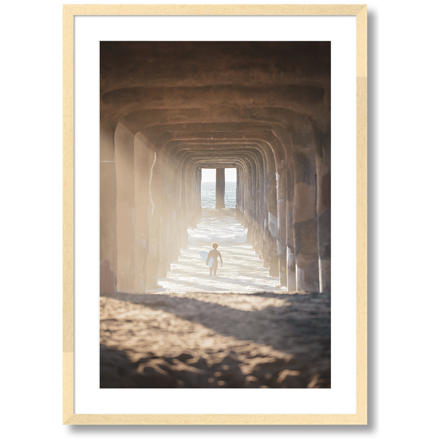 image of a surfer under a pier walking out into the waves carrying a surfboard