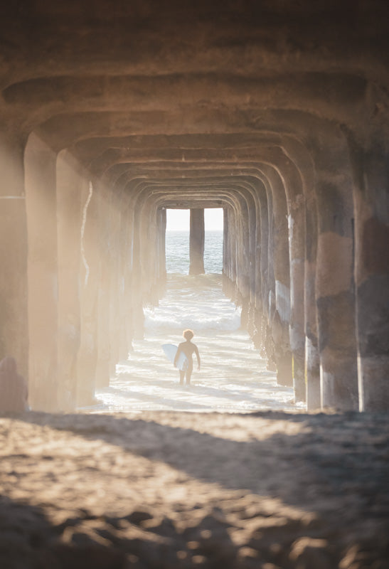 image of a surfer under a pier walking out into the waves carrying a surfboard